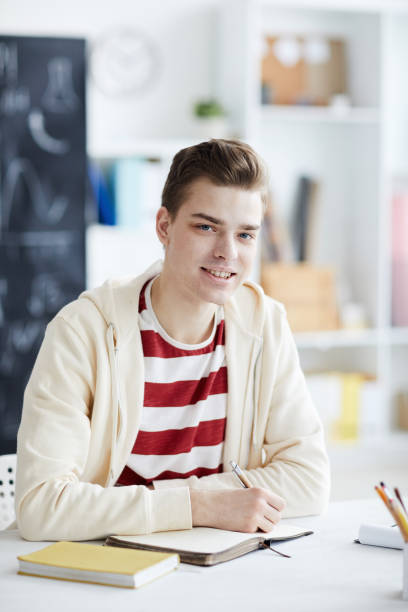 istock Smiling handsome young student in hoodie sitting at table in classroom and making notes in organizer while looking at camera 1132662698
