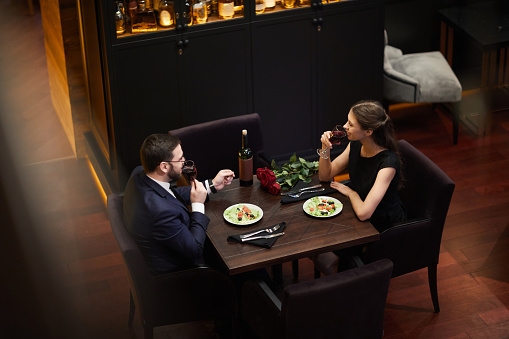 Young amorous dates sitting by table in luxurious restaurant, drinking red wine and talking before eating dinner