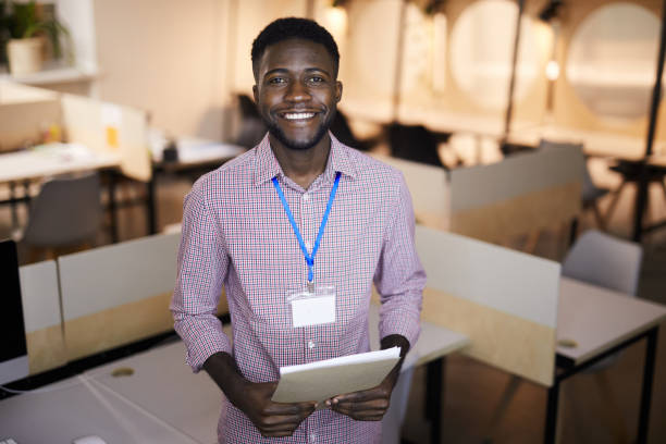 Cheerful excited handsome young black guy with stubble wearing blank badge holding notepad and looking at camera while standing in coworking space
