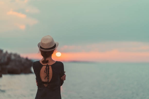 Woman enjoying the sunset on beach holiday Rear view of woman with sun hat staring the sunset over the water depression land feature stock pictures, royalty-free photos & images