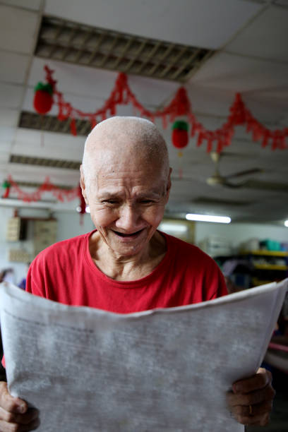 A senior man holding a copy of newspaper and reading with cheerful expression.