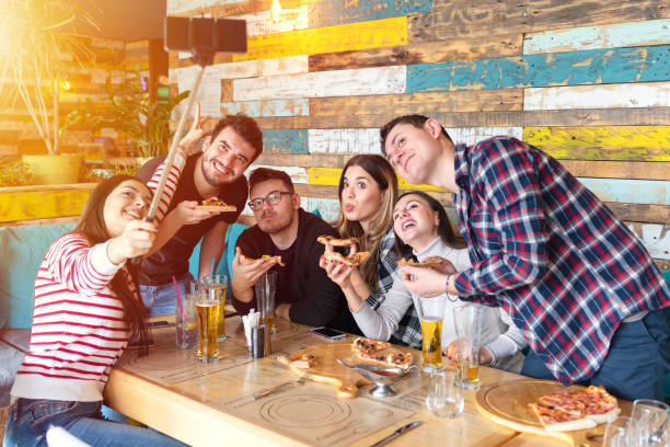 Cheerful young friends taking selfie with smart phone while sharing pizza at pub restaurant Cheerful young friends taking selfie with smart phone while sharing a pizza, Young people at table in restaurant using mobile phone to take selfie while eating and share on social media friends in bar with phones stock pictures, royalty-free photos & images