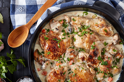 chicken fricassee in a black dutch oven - chicken meat browned and braised in white wine creamy sauce with mushrooms and vegetables, classic french recipe, view from above, flat lay, close-up