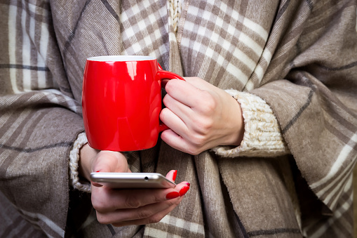 Hipster blogger with red cup coffee in hands reading incoming sms message on smartphone connected to wifi. Young student watching broadcasting online on modern mobile phone