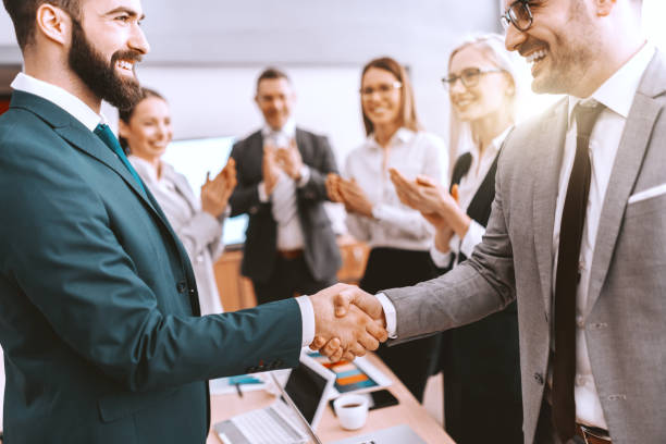 istock Two partners shaking hands while other colleagues clapping hands. We grow by lifting others. 1132730600
