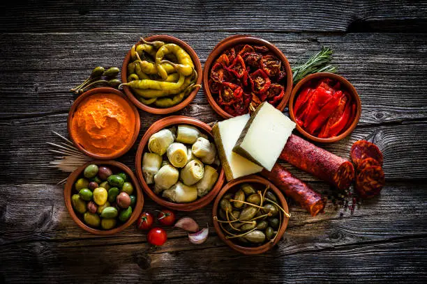 Spanish food: tapas. Chorizo, manchego cheese, olives, capers, jalapeño peppers, pimientos piquillo, artichoke hearts and sun dried tomatoes shot from above on dark rustic wooden table. The food is in clay bowls. Predominant colors are red and brown. Low key DSRL studio photo taken with Canon EOS 5D Mk II and Canon EF 70-200mm f/2.8L IS II USM Telephoto Zoom Lens