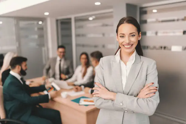 Photo of Beautiful Caucasian brunette in formal wear standing with arms crossed and smiling. In background colleagues having meeting. You don't build a business. You build people and then people build the business.