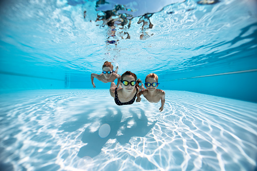 Three cute happy kids swimming underwater in the huge swimming pool towards the camera. Shot with Nikkor 14-24 on D850. Wide shot with plenty of copy space and multiple crops possible.\nPerfect sharpness and huge resolution.