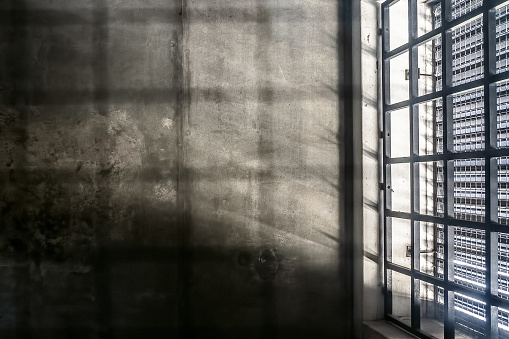 The very sober interior of a prison cell: barred windows with little light coming in and bare concrete walls