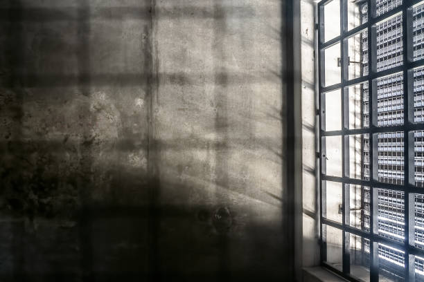 the very sober interior of a prison cell: barred windows with little light coming in and bare concrete walls - cela imagens e fotografias de stock