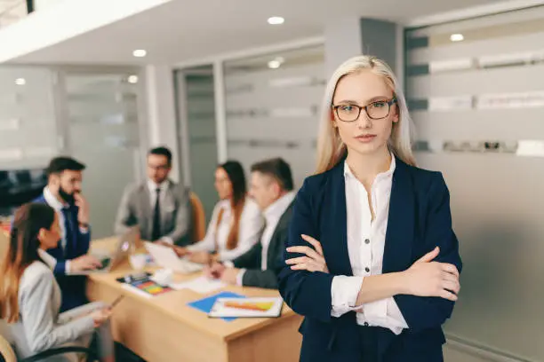 Photo of Portrait of attractive blonde businesswoman in formal wear holding crossed arms while standing in board room In background colleagues having meeting. Be somebody who makes everybody feel lime somebody.