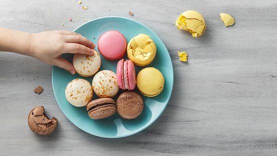 Little, kid's hand reaching for a macaroon from a turquoise plate on a wooden textured table. Copy space