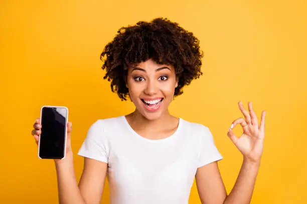 Close-up portrait of her she nice cute attractive charming cheerful cheery ecstatic optimistic wavy-haired lady holding in hands cell showing ok-sign isolated on bright vivid shine yellow background.