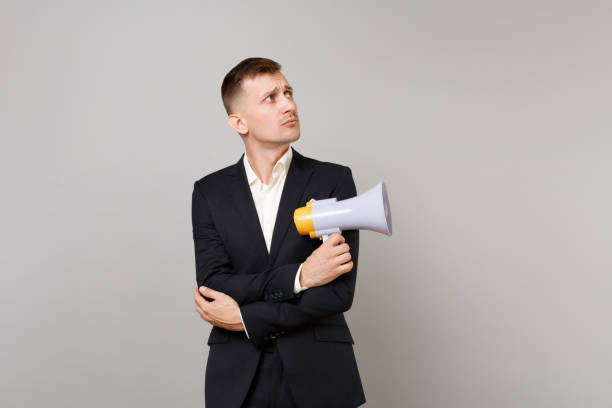 Young business man in classic black suit shirt looking aside holding hands with megaphone crossed isolated on grey background in studio. Achievement career wealth business concept. Mock up copy space joven empresario en camisa de traje negro clásico mirando a un lado sosteniendo las manos con megátero cruzado aislado sobre fondo gris en estudio. concepto del negocio de la abundancia de la carrera del logro. maqueta de espacio de copia.