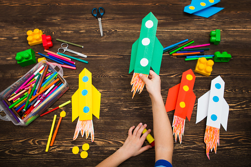 the child holds the rocket made of paper against the background of a wooden table. preschool Child in creativity in the home. Happy kid makes rockets from paper. Children's creativity.