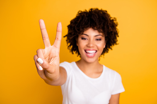 Close up photo beautiful amazed she her dark skin lady glad arms hands fingers raised show v-sign peaceful hippie concept wearing casual white t-shirt isolated yellow bright background.