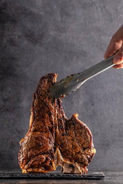 Large beef roast T-bone steak with smoke. Head chef holding steak meat tongs on a black background. photon image. copy space Large beef roast T-bone steak with smoke. Head chef holding steak meat tongs on a black background. photon image. copy space fire alphabet letter t stock pictures, royalty-free photos & images