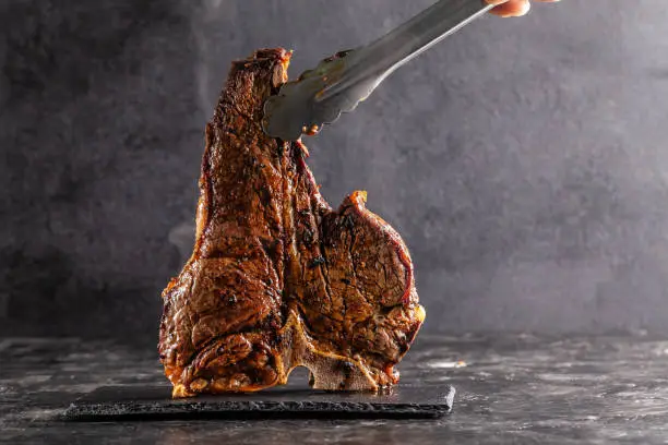 Photo of Large beef roast T-bone steak with smoke. Head chef holding steak meat tongs on a black background. photon image. copy space