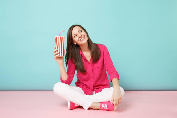 Full length portrait woman in rose shirt blouse, white pants sitting on floor hold cup of soda isolated on pink blue pastel wall background studio. Fashion lifestyle concept. Mock up copy space mujer de retrato de longitud completa en camisa de rosa blusa pantalones blancos sentados en el suelo mantener la taza de soda aislada en el estudio de fondo de pared azul pastel rosa brillante. concepto de estilo de vida de moda. maqueta de espacio de cop