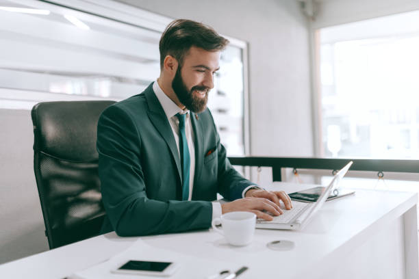 homme d'affaires barbu heureux caucasien dans l'usure formelle utilisant l'ordinateur portatif dans le bureau. un vainqueur est un rêveur qui ne quitte jamais. - technology contemplation clothing formalwear photos et images de collection