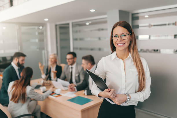 porträt der wunderschönen geschäftsfrau in formaler kleidung, mit langen braunen haaren und brillen mit zwischenablage im boardzimmer. im hintergrund diskutieren coworker. streben sie nach fortschritt, nicht nach perfektion. - success practicing leadership leading stock-fotos und bilder