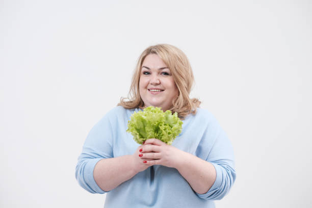 une jeune femme grasse luxuriante dans les vêtements bleus occasionnels sur un fond blanc retenant la laitue verte laisse dans ses mains et la bouche. - overweight women salad frustration photos et images de collection