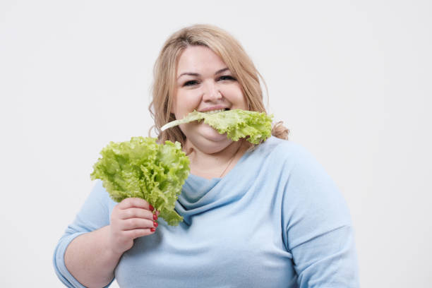 une jeune femme grasse luxuriante dans les vêtements bleus occasionnels sur un fond blanc retenant la laitue verte laisse dans ses mains et la bouche. - overweight women salad frustration photos et images de collection