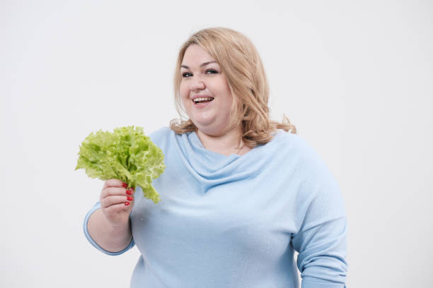 une jeune femme grasse luxuriante dans les vêtements bleus occasionnels sur un fond blanc retenant la laitue verte laisse dans ses mains et la bouche. - overweight women salad frustration photos et images de collection