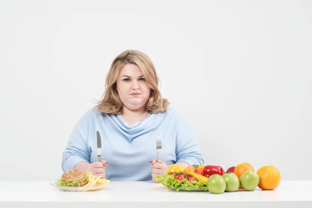 jeune femme de graisse sinueuse dans les vêtements bleus occasionnels sur un fond blanc à la table choisit ce qu'il faut manger: fast-food ou aliments sains. alimentation et bonne nutrition. - freedom sandwich bread food photos et images de collection