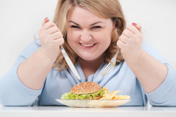 jeune femme grasse sinueuse dans des vêtements bleus occasionnels sur un fond blanc à la table mangeant avec impatience la restauration rapide, hamburger et français frites. alimentation et bonne nutrition. - freedom sandwich bread food photos et images de collection