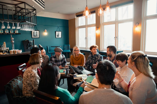 Enjoying a Drink at Book Club Large group of people with a mixed age range sitting together around a table. They are enjoying a drink and having a book club meeting. casual clothing stock pictures, royalty-free photos & images