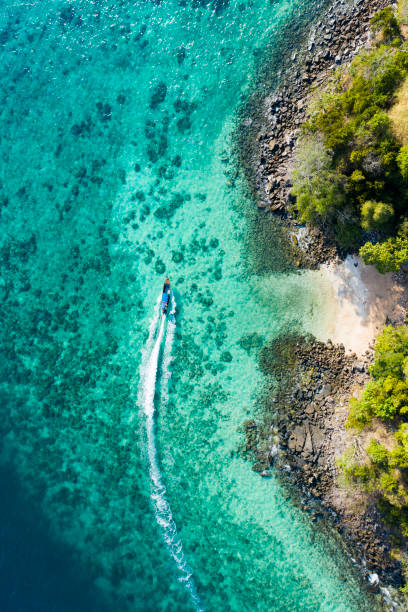 View from above, aerial view of a traditional long-tail boat sailing near a stunning barrier reef with a beautiful small beach bathed by a transparent and turquoise sea. Phi Phi Island, Thailand. View from above, aerial view of a traditional long-tail boat sailing near a stunning barrier reef with a beautiful small beach bathed by a transparent and turquoise sea. Phi Phi Island, Thailand. satun province stock pictures, royalty-free photos & images