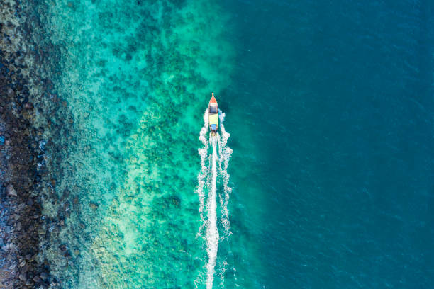 View from above, aerial view of a traditional long-tail boat sailing near a stunning barrier reef with a beautiful small beach bathed by a transparent and turquoise sea. Phi Phi Island, Thailand. View from above, aerial view of a traditional long-tail boat sailing near a stunning barrier reef with a beautiful small beach bathed by a transparent and turquoise sea. Phi Phi Island, Thailand. satun province stock pictures, royalty-free photos & images
