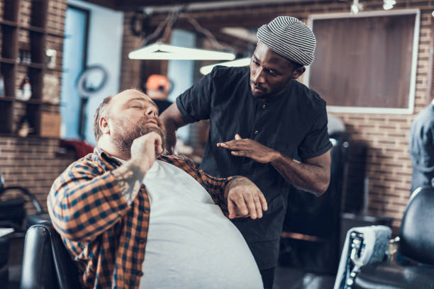 Two person in barber shop speaking about beard cut Handsome client sitting in comfort chair indoor barbershop. He explains his wishes about beard cut to master black barber stock pictures, royalty-free photos & images