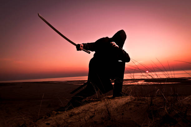 istock Assassin in black suit with sword working out at the beach. 1132790653