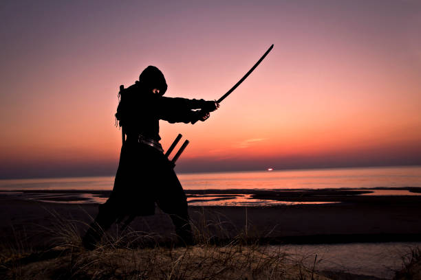 istock Assassin in black suit with sword working out at the beach. 1132791366