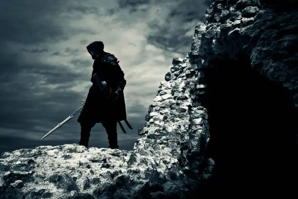 Warrior in black suit and with sword is standing next to the ruined fortress. Backside view. Cloudy sky.