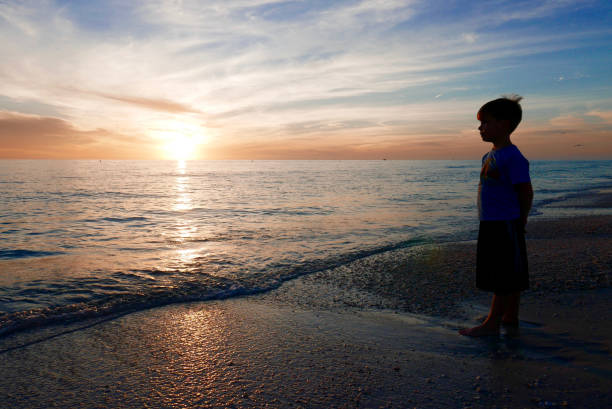 Silhouette of a young boy standing on a Florida gulf coast beach at the water's edge, looking at the sunset in the evening