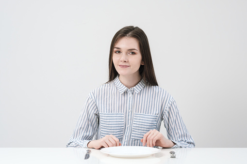Hungry thin girl sitting at the table in front of an empty plate with a knife and fork. Concept of diet and hunger.