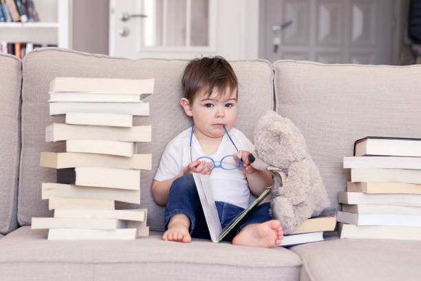 niedlicher, kluger, lustiger kleiner junge mit nachdenklichem ernstem gesichtsausdruck hält gläser in den händen lesen buch sitzen auf dem sofa mit teddy-bär-spielzeug und haufen von büchern zu hause mit blick auf die kamera. kindererziehung und-entwi - furniture barefoot little boys family stock-fotos und bilder