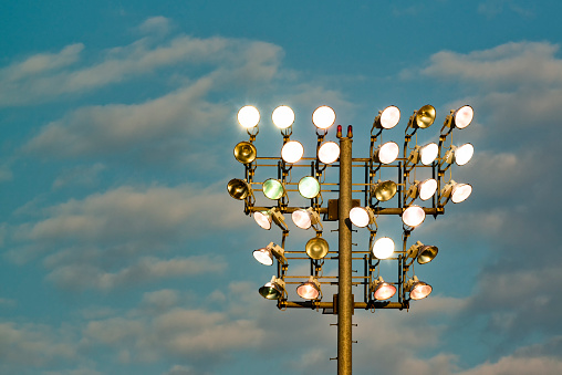 A group of high intensity lamps to illuminate a sports field atop a steel pole as the sun sets with clouds in the background