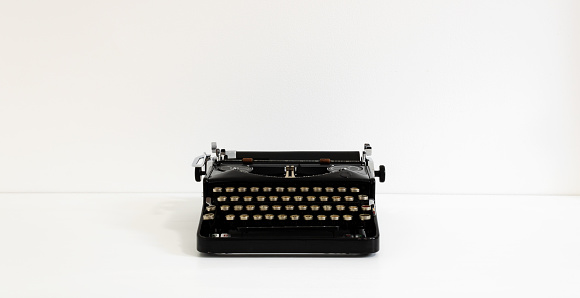 Old retro vintage black typewriter in black color laid on white desk in front of white wall with copy space