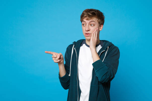 istock Perplexed disgusted young man in casual clothes putting hand on cheek and pointing index finger aside isolated on blue wall background. People sincere emotions, lifestyle concept. Mock up copy space. 1132857295