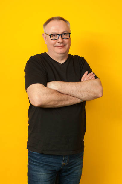 portrait de studio de 50 ans homme de calvitie en lunettes, jeans et t-shirt noir sur fond jaune - 45 49 years photos et images de collection