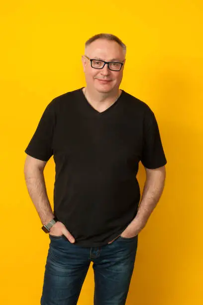 Studio portrait of 50 years old balding man in glasses, jeans and black t-shirt on yellow background