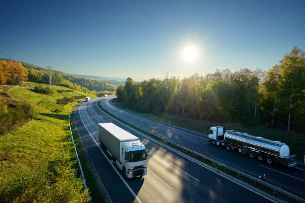 istock Three white trucks and chrome tanker driving on the asphalt highway between deciduous forest under the rays of the sun. View from above. 1132878295