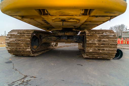 Bottom of an excavator with soil on grouser pad. The dirty bottom part of a yellow excavator with soil on the grouser pads. Trees, homes, construction cones, and sky can be seen behind the vehicle.