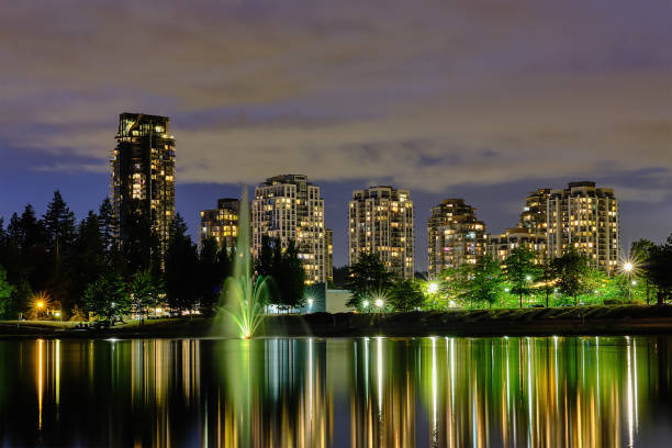 Nightscape city view, Coquitlam, Greater Vancouver area, Canada. Fountain in lake and water reflections of modern buildings windows lights