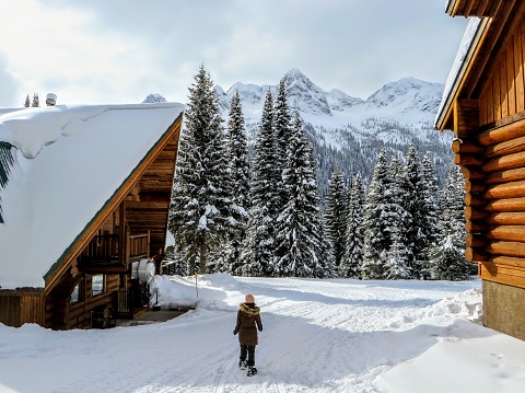 A young female snowshoer walking in between two wood cabins during the winter in the remote snow covered forests of Fernie, British Columbia, Canada.