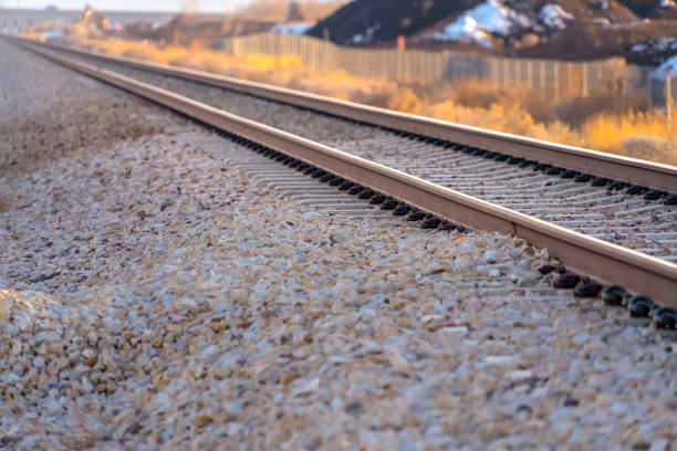 Railroad track on a dry and rocky ground. Close up view of a railroad track over a rocky terrain on a bright and sunny day. Sunlit shrubs and buildings can be seen in the blurry background.